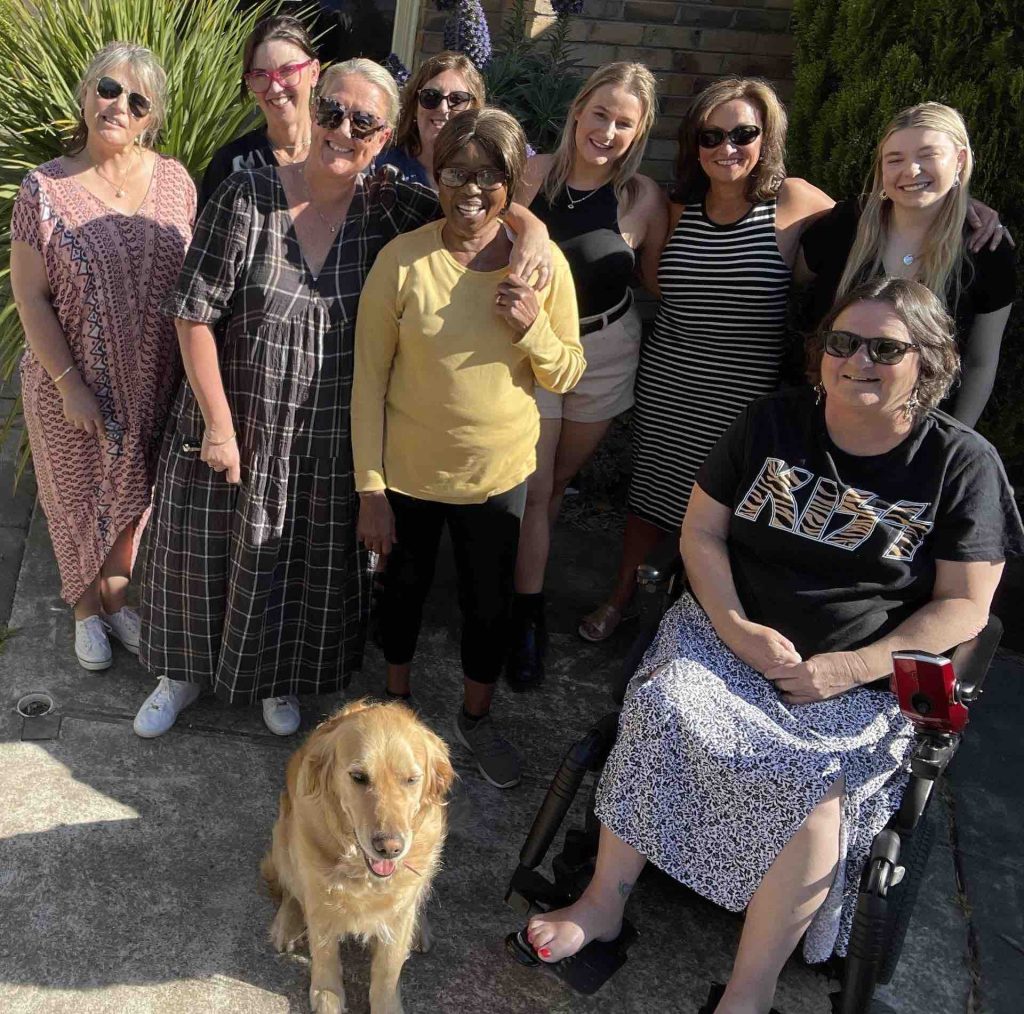 A smiling group of Destination Accessible guests and team members outdoors, including a woman using a power wheelchair, with a golden retriever in front
